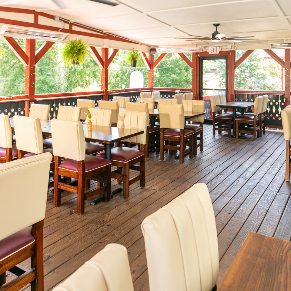 Outdoor restaurant patio with wooden floors, red beams, cream and maroon chairs, and hanging plants.