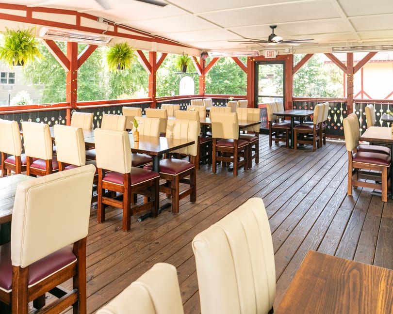 Outdoor restaurant patio with wooden floors, red beams, cream and maroon chairs, and hanging plants.