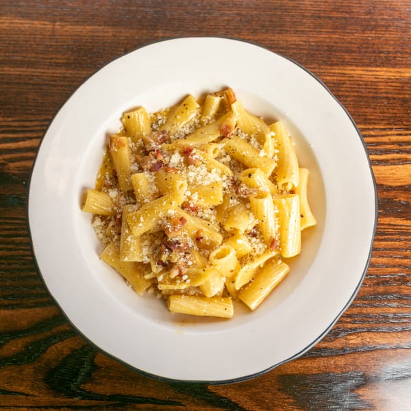 A white bowl of rigatoni pasta with a creamy sauce, bacon bits, and grated cheese on a wooden table.