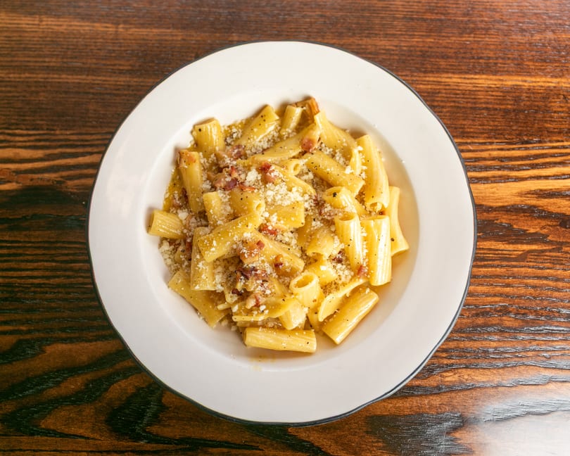 A white bowl of rigatoni pasta with a creamy sauce, bacon bits, and grated cheese on a wooden table.