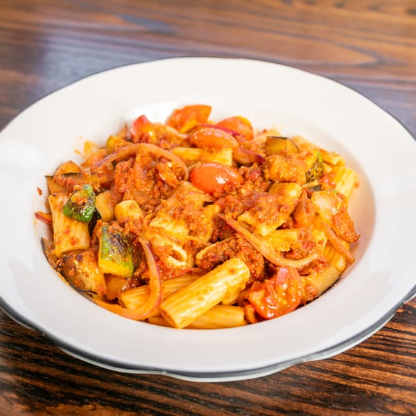 Rigatoni pasta with a rich red sauce, cherry tomatoes, zucchini, and red onions in a white bowl.