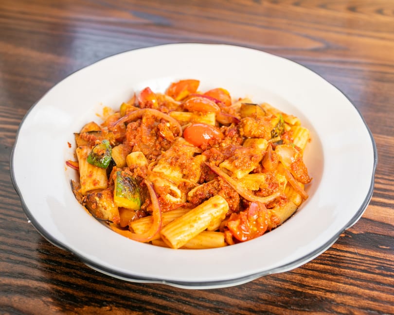 Rigatoni pasta with a rich red sauce, cherry tomatoes, zucchini, and red onions in a white bowl.