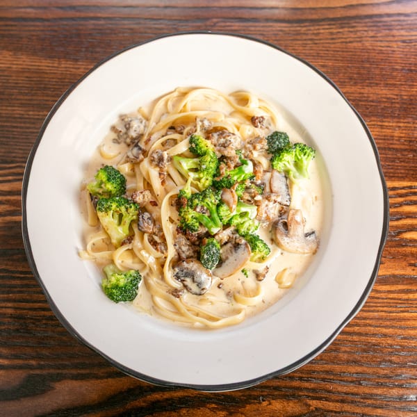 Creamy fettuccine pasta with broccoli, mushrooms, and ground meat in a white bowl on a wooden table.