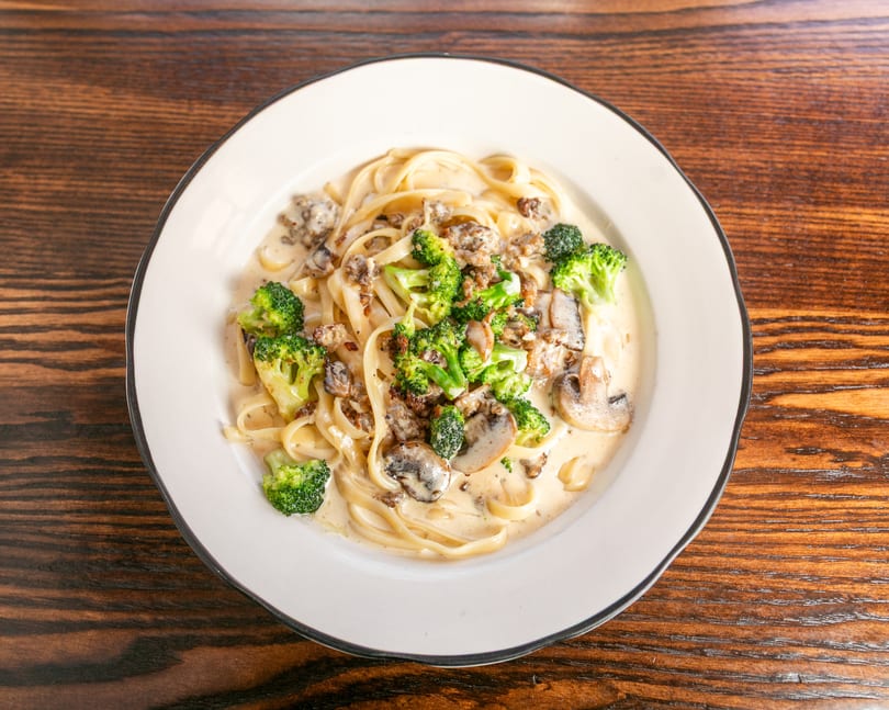 Creamy fettuccine pasta with broccoli, mushrooms, and ground meat in a white bowl on a wooden table.