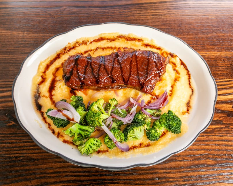Grilled steak with balsamic glaze on a bed of polenta, garnished with broccoli and red onions.
