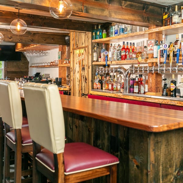 Rustic bar with wooden beams, shelves of liquor bottles, beer taps, and cream-colored bar stools.