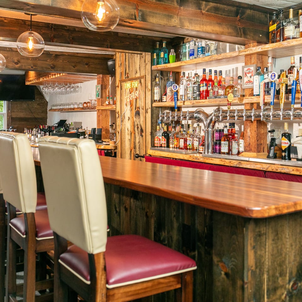 Rustic bar with wooden beams, shelves of liquor bottles, beer taps, and cream-colored bar stools.