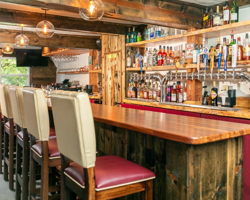 Rustic bar with wooden beams, shelves of liquor bottles, beer taps, and cream-colored bar stools.