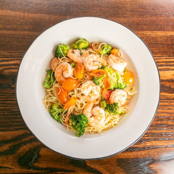 Shrimp scampi with angel hair pasta, broccoli, and cherry tomatoes in a white bowl on a wooden table.