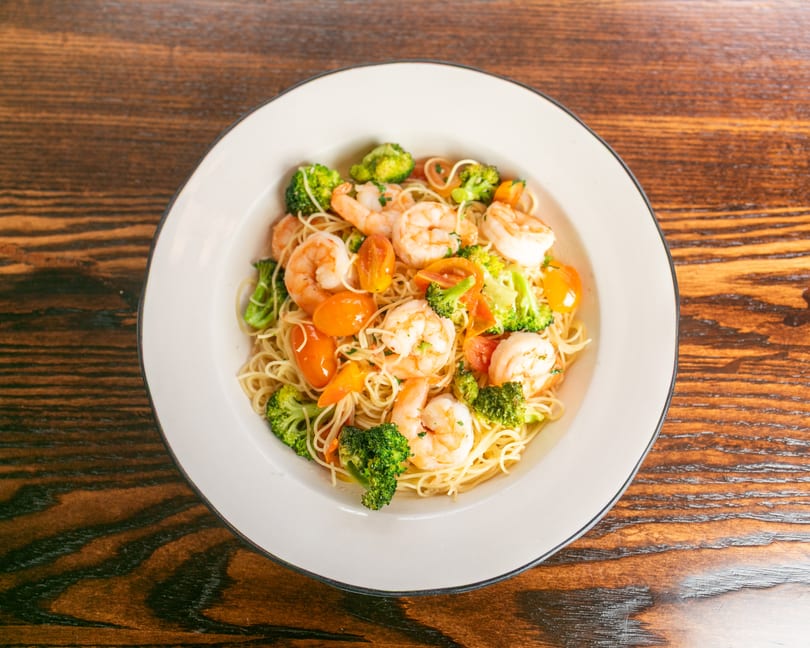 Shrimp scampi with angel hair pasta, broccoli, and cherry tomatoes in a white bowl on a wooden table.
