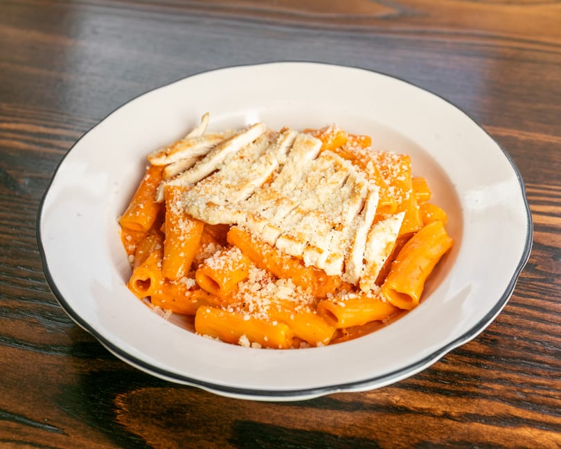 Rigatoni pasta with orange sauce, grilled chicken, and grated cheese in a white bowl.